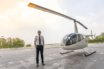 Picture of a male helicopter pilot in his white shirt with tie uniform. He is prepared for his flight. He stands in front of a white helicopter in the open area prepare to take off