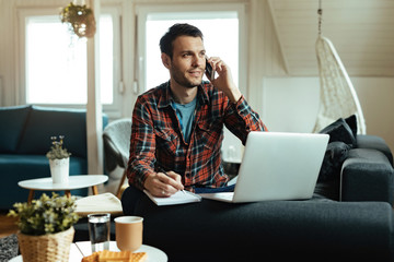 Smiling man talking on the phone while taking notes and working at home.