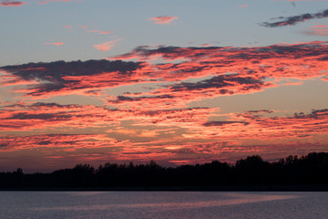 Vibrant colorful sunset by the water / pink orange red clouds burning with colors / lake landscape sunrise / horizon trees and forest