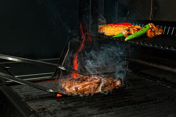 Beef steak and vegetables are grilled and turned over with tongs. The photo shows flames and smoke....