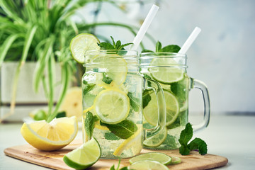 Two glasses of homemade lemon, lime, and mint lemonade sit on the wooden dining table. Cold, refreshing summer lemonade or mojito. - obrazy, fototapety, plakaty