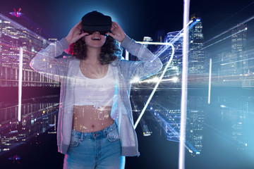 Teenage curly girl is playing in game club. Smiling surprised young woman in VR glasses is watching 3d virtual city tour in simulator. Entertainment and leisure concept. Modern technologies.