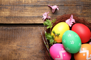 Obraz na płótnie Canvas Colorful Easter eggs in decorative nest with flowers on wooden background, flat lay. Space for text