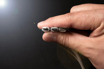 Hand is holding set of smartphone connectors: lightning, micro-usb and type-c cables on dark background.