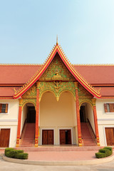 An entrance of Buddhist temple with ornate red roof with golden decoration in Vientiane in Laos against blue sky