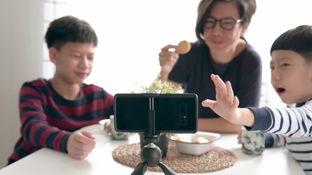 Asian mother and her two boys make online video call to father, show their biscuits, wave hands at afternoon snack time at home due to social distancing and school closure during Covid-19 pandemic