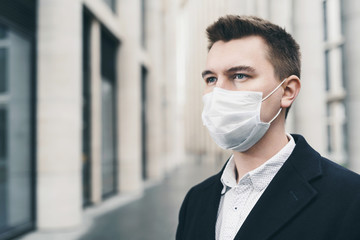 Businessman and coronavirus epidemic. Close up portrait of young business man in a disposable facial mask. A man defends himself against covid 19 on the big city street. Modern buildings at background