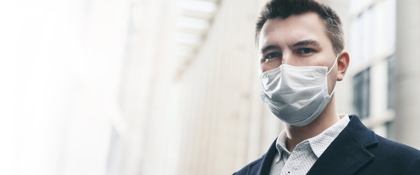 Businessman And Coronavirus Epidemic. Close Up Portrait Of Young Business Man In A Disposable Facial Mask. A Man Defends Himself Against Covid 19 On The Big City Street. Modern Buildings At Background