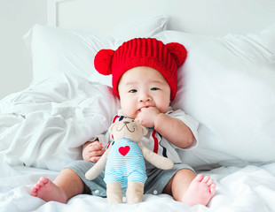 Portrait of a newborn Asian baby boy, Charming child 5 month old wore a suit and a red wool hat sitting in bedroom sucking his finger,fat baby cute and smilingly with a doll,soft selective focus