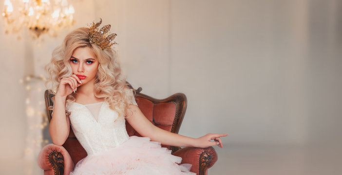 Portrait capricious princess girl. Blond hair gold crown. lady woman sitting in vintage armchair. backdrop white room lamp chandelier. Bored queen hand shows gesture on free space for text wide screen
