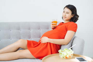 Obraz na płótnie Canvas Pretty young Asian pregnant woman eating salad and drinking milk when sitting on sofa.