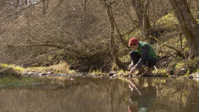 Young handsome man washes the espresso maker and cup in a forest river. Bright and sunny early sping day. Cold weather. Wide shot