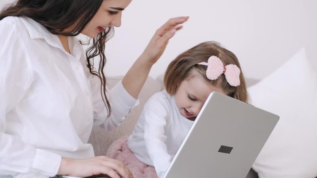 The pretty mother and her child girl are studying with a laptop. The mother is teaching heer kid. The girl is getting along with the parent typing on the keyboard and watching something on hte laptop