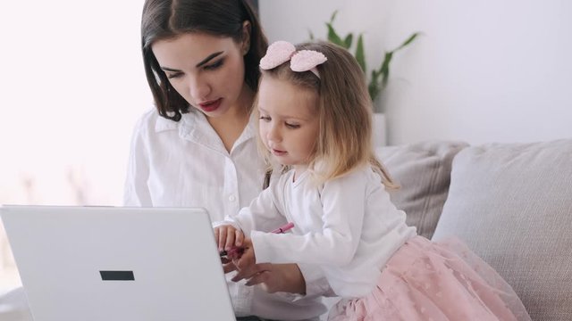 The pretty mother and her child girl are studying with a laptop. The mother is teaching heer kid. The girl is getting along with the parent typing on the keyboard and watching something on hte laptop