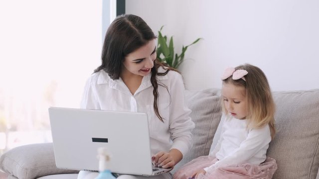 The pretty mother and her child girl are studying with a laptop. The mother is teaching heer kid. The girl is getting along with the parent typing on the keyboard and watching something on hte laptop