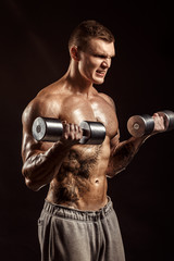 Serious tattoed shirtless athlete lifting metal dumbbells training on dark background.