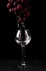 a glass glass stands on the table against a black background. A branch of red grapes hangs from above. Water drops from the top of the grapes into the glass
