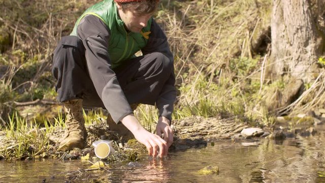Young handsome man washes the espresso maker and cup in a forest river. Bright and sunny early sping day. Cold weather