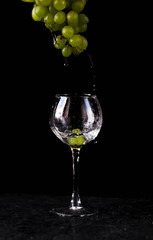 a glass glass stands on the table against a black background. A branch of green grapes hangs from above. Water drops from the top of the grapes into the glass