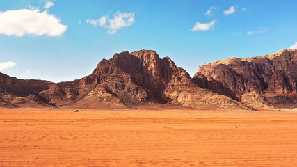 Flat red desert with large mountains in distance, blue sky above, some off road vehicles in background, typical scenery at Wadi Rum, Jordan