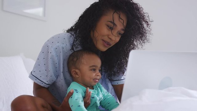 Mother Wearing Pyjamas Cuddling Baby Son In Bedroom Whilst Working From Home On Laptop