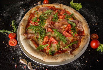 Pizza with prosciutto, arugula and chopped tomatoes, topped with cheese. On a black table with sprinkled spices and ingredients. Photo for the menu.