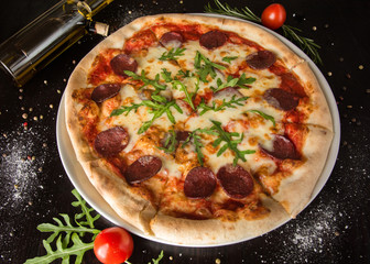 Sudzhuk pizza - with chopped salami, arugula and cheese. On a black table with sprinkled spices and ingredients. Photo for the menu.