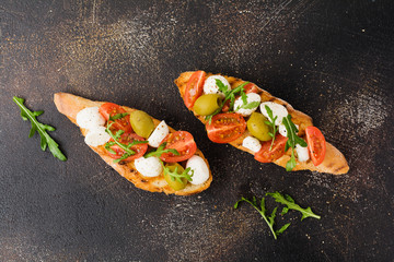 Caprese bruschetta toasts with cherry tomatoes, mozzarella, olives and basil on old dark background. Top view.
