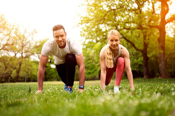 Naklejka na ściany i meble Fit young man and woman exercising in park. Smiling caucasian couple doing workouts on grass on sunny morning.