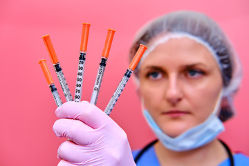 Portrait of a doctor woman in a medical beret with syringes in her hands, closeup. Nurse in a blue uniform and a protective mask on a pink background.