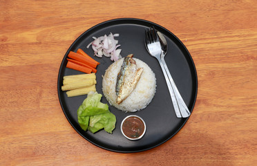 Top view breakfast in the black round plate on wooden floor. Fried mackerel and Rice with Spicy Shrimp Paste Dip and vegetable, Chinese cabbage, baby corn, carrots and sliced onions.