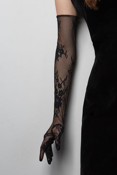 Cropped shot of a lady in a black tight dress and a black evening  glove against the gray wall. The elegant elbow glove is made of the lace fabric with floral pattern. 
