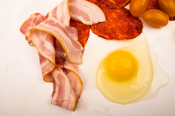 A broken chicken egg without a shell, slices of sausage and bacon and tomatoes on a white background. Close up.