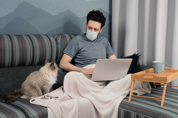 A man in a medical mask is working on a laptop sitting at home on the sofa and a cat is sitting next to him