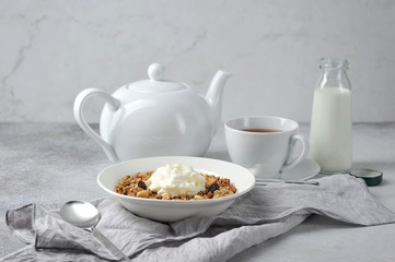Table setting for breakfast.  Plate with granola and yogurt.  A teapot, a cup with tea and a bottle with milk in the background.  Light background.