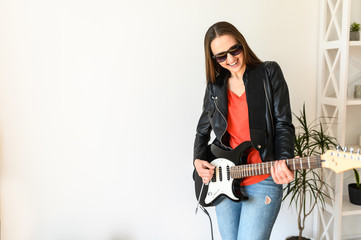 Home leisure with fun. A young beautiful woman in sunglasses and in leather jacket is playing an electric guitar indoors, she is smiling happily. Hobby concept, music talent