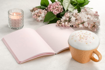 Pink notebook on the table with a large cup of cappuccino, a candle and a bouquet of lilac