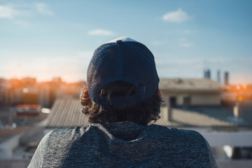 Photo taken from behind of a boy in a capwith long hair. look at the city from the roof of a...