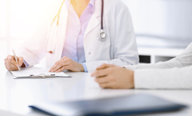 Unknown woman-doctor and female patient sitting and talking at medical examination in clinic, close-up. Therapist wearing blue blouse is filling up medication history record. Medicine concept