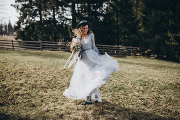 Portrait of the bride. Country style. A young girl in a blue gray wedding dress, denim jacket and hat with a bouquet of flowers and greenery in her hands on a background of a forest and a wooden fence