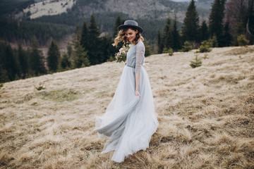 Portrait of the bride. A young girl in a blue gray wedding dress and hat with a bouquet of flowers and greenery in her hands on a background of mountains and a forest at sunset