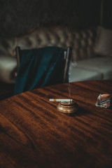 A smoldering burning cigarette on golden ashtray on a wooden table. White curly smoke on a room background with vintage furniture, chair and sofa. No smoking concept. Artwork decoration with cigarette