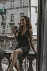 A happy young beautiful smiling caucasian woman sitting on a rail at vintage balcony and holding a retro film camera. A pretty tanned girl with curvy long brown hair taking pictures, view from bedroom
