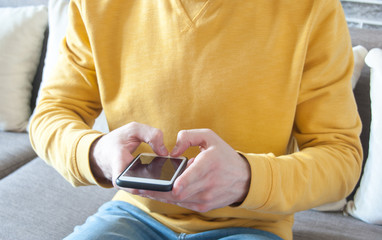 Professional freelancer man or student or teenager in a yellow sweater sitting on the sofa at home and using his smartphone. Freelancer concept. Studying concept. Work from home concept. Chatting