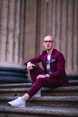 Obraz premium A young bald guy with glasses, stylishly dressed in a purple suit sitting on stairs next to the columns