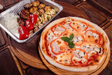 Roasted potatoes with grilled vegetables and meat on baking tray and pizza on wooden background