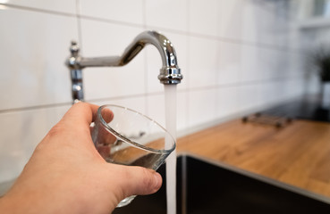 mans hand holding an empty glass nearby a stainless steel kitchen tap. person consider pouring running water from chrome faucet. Safe to drink or not fit for human consumption. Potable or Non-potable