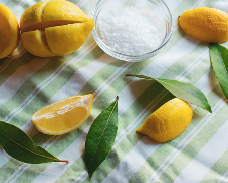 An arrangements of lemons, sea salt and bay leaves on a green and white striped background.  The lemons are in slices, whole and being prepared to make preserved lemons