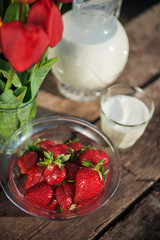 Fresh strawberries on plate on old wooden background. Red tulips in vase on table. Jar and glass of milk. Breakfast time. Healthy food. Vegetarian dish. Spring time
