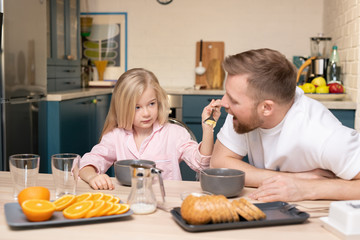 Adorable little girl holding spoon with corn flakes by her father mouth by table